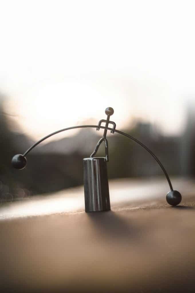 Metal wire sculpture of a small stick-figure balancing on a black cylindrical base, with two curved arms ending in spherical weights on a wooden surface at sunset.