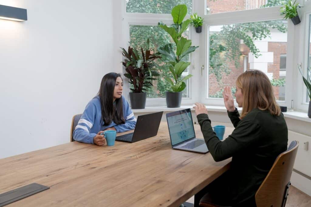 Two women sit across from each other at a wooden table with laptops, mug cups, and a bright plant-filled office as they discuss and gesture during a meeting.