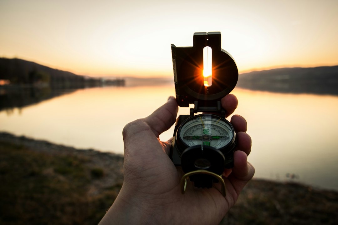 Hand holding a compass at sunset beside a calm lake, sun peeking through the device's lens shield