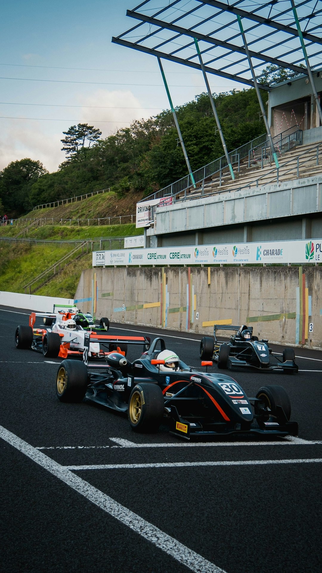 Open-wheel race cars on a dark asphalt track near a concrete wall with sponsor banners, stadium seating, and a grassy hill in the background.