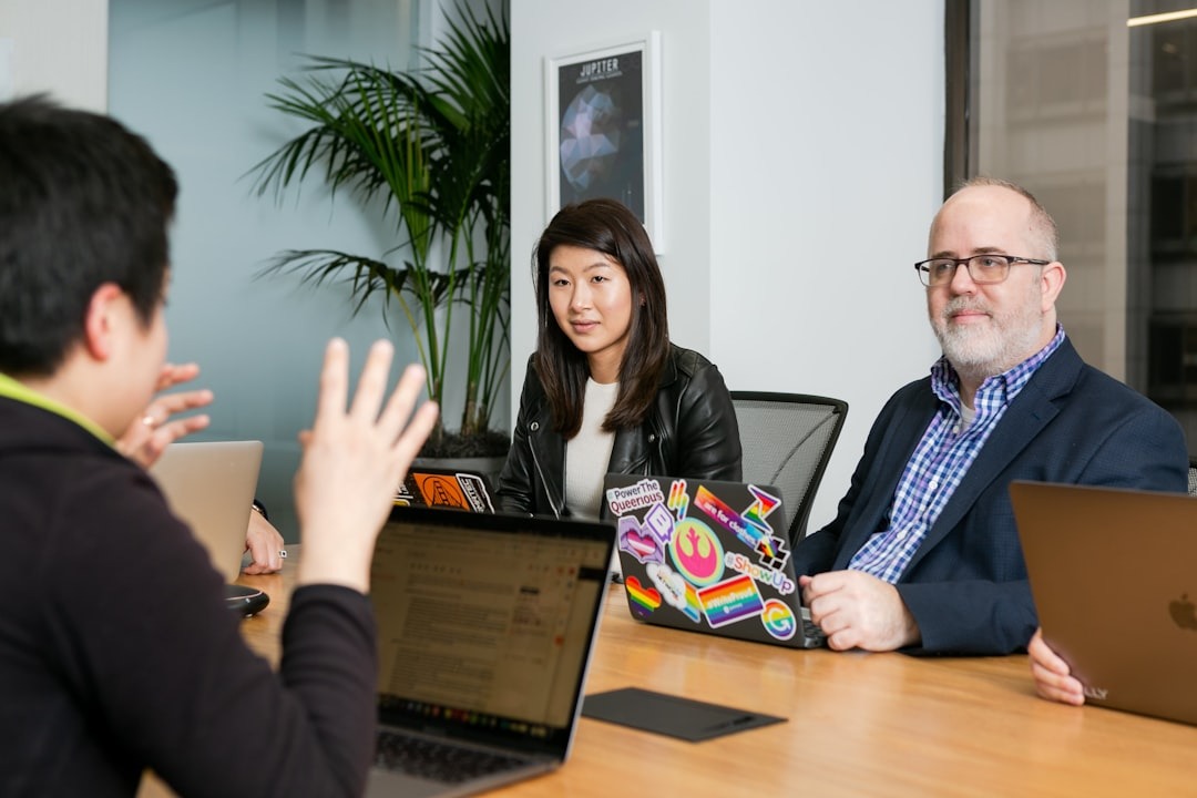 Three coworkers sit around a wooden conference table with laptops, one person gesturing with raised hands.