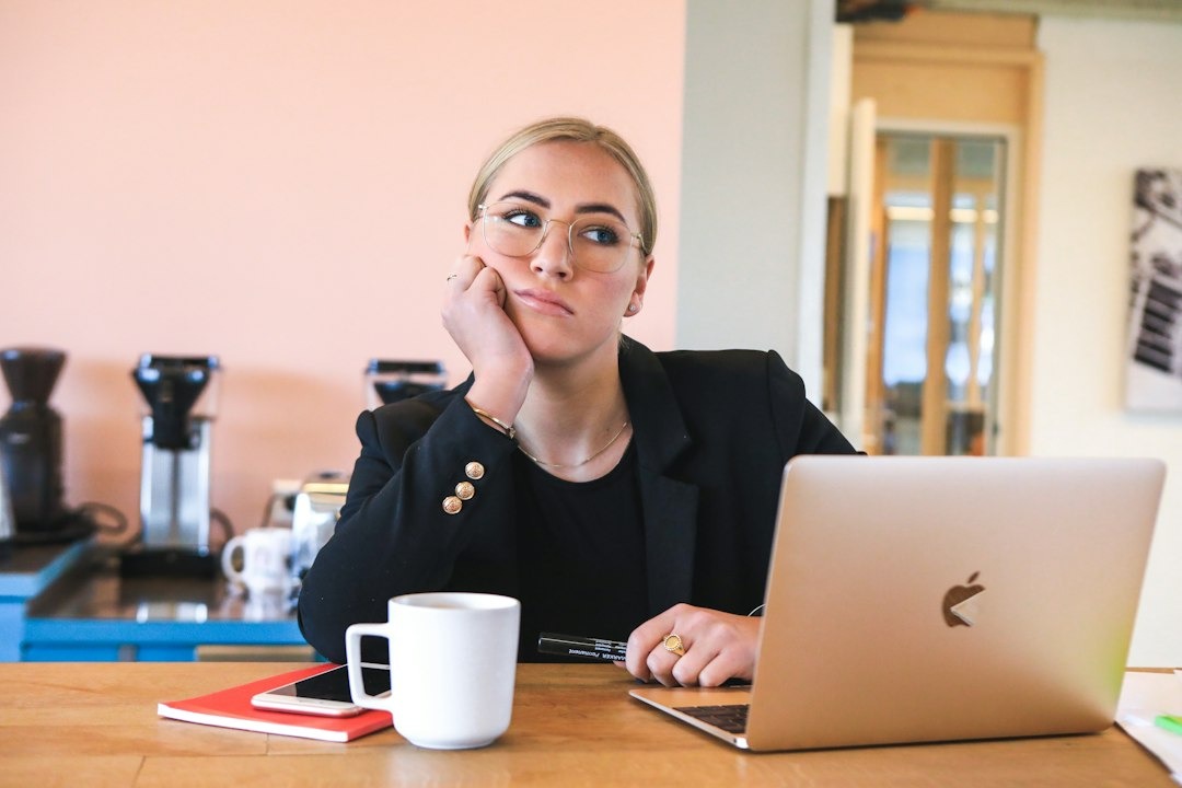 Professional woman in a blazer and glasses sits at a wooden desk with a MacBook, resting her chin on her hand.