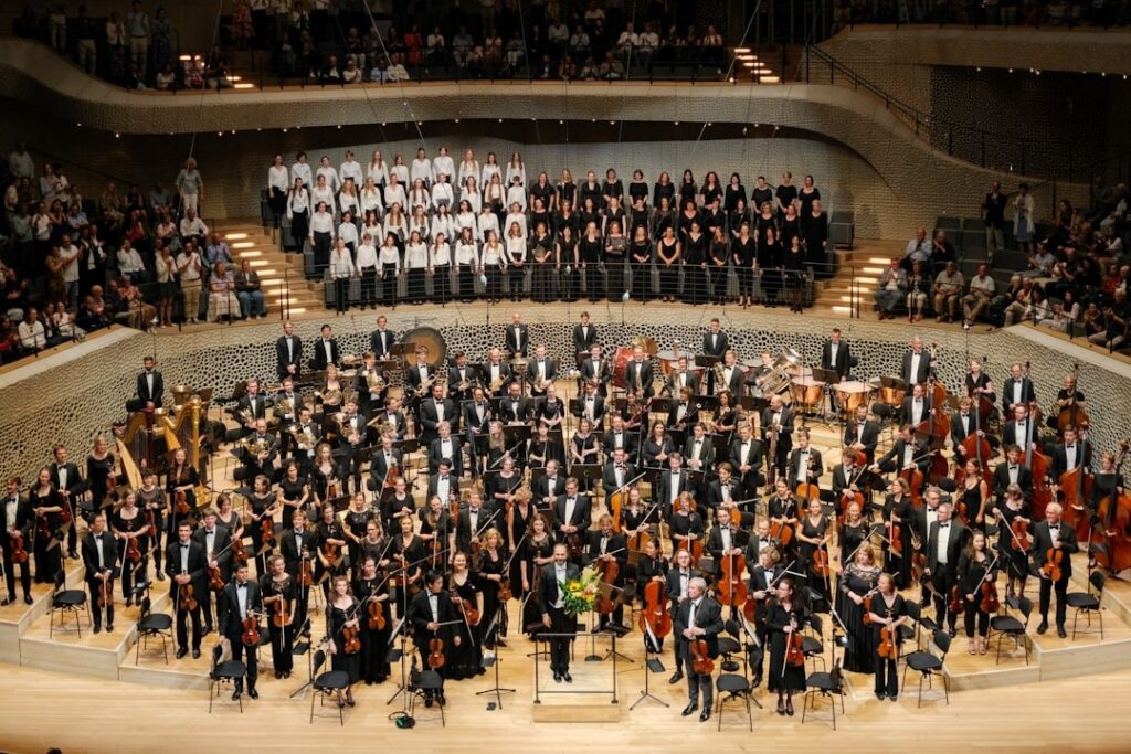 Full symphony orchestra on stage in a concert hall, conductor at the podium, with a large mixed choir on a balcony above and audience in the seats behind them.