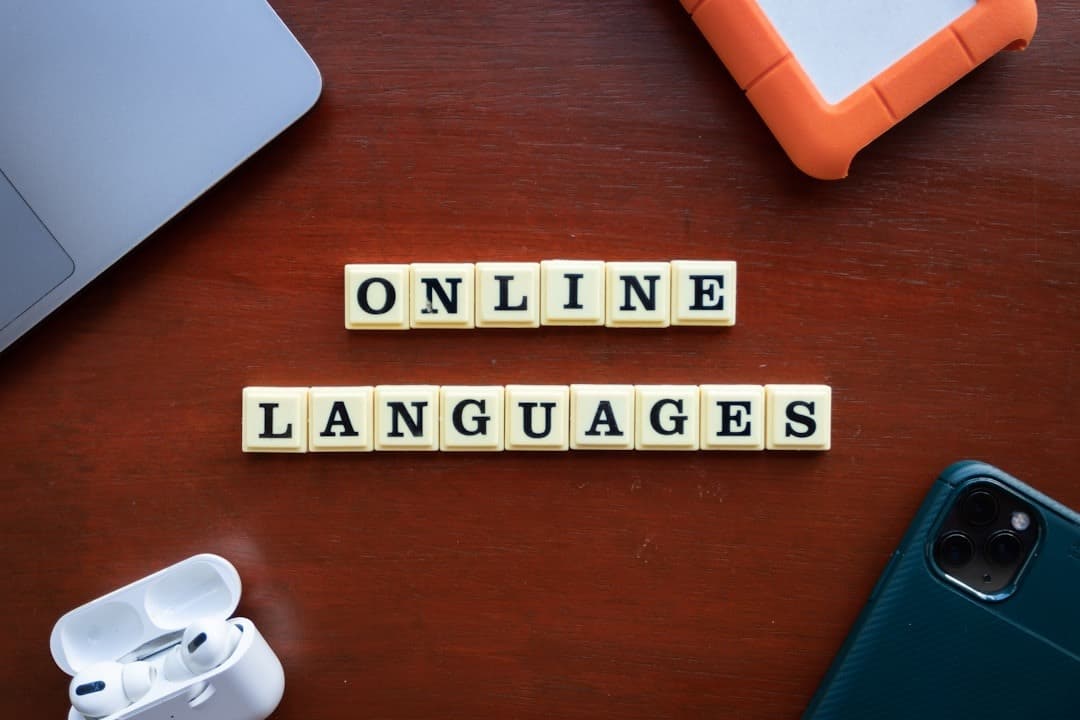 Scrabble tiles spell 'ONLINE' over 'LANGUAGES' on a wooden desk with a laptop, earbuds, phone, and a protective case nearby.