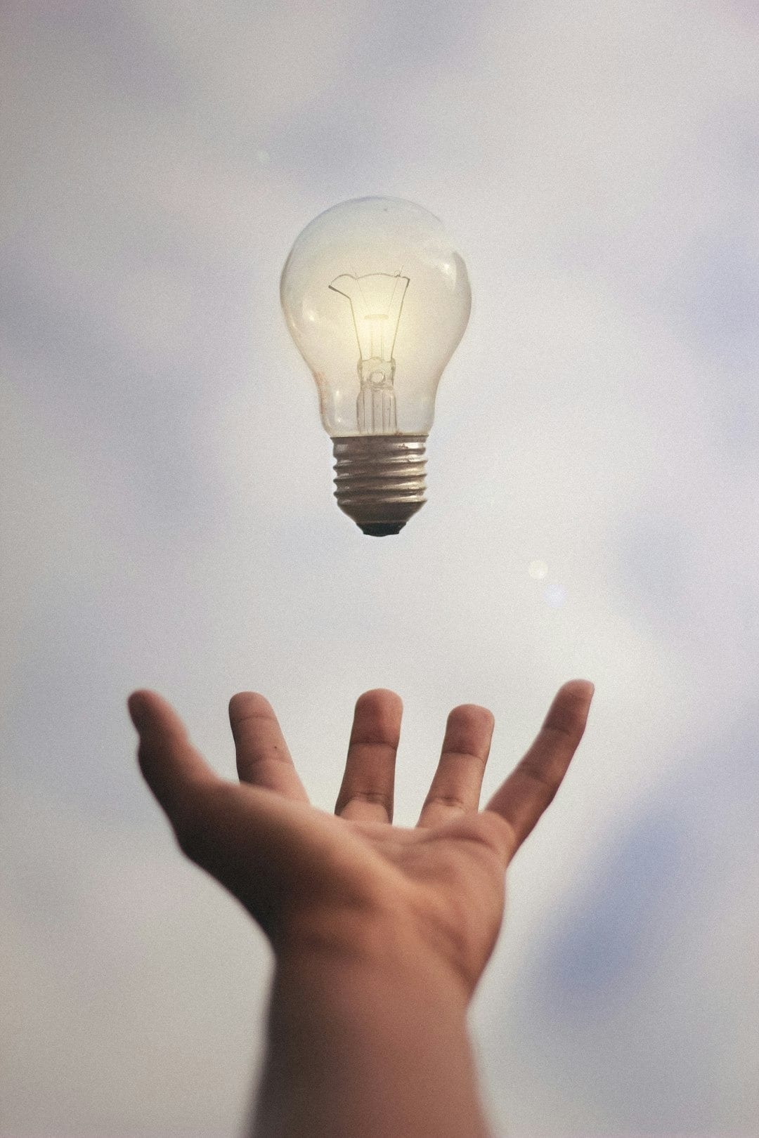 Hand reaching up toward a floating light bulb against a pale sky, as if catching a spark of an idea.