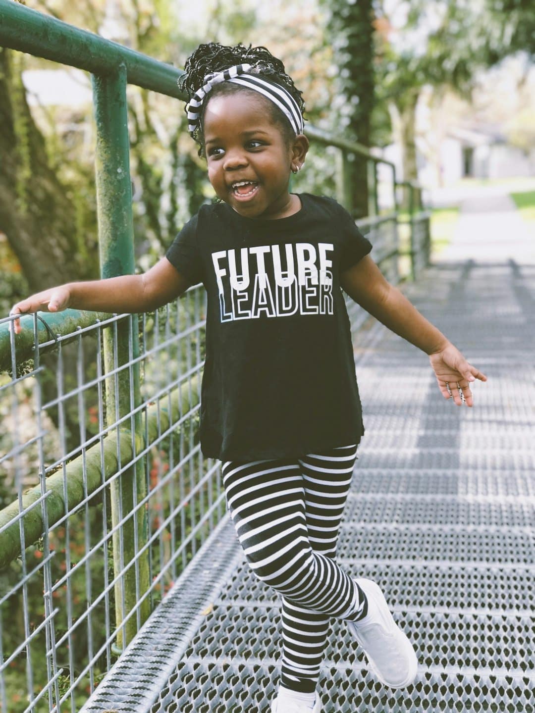 Young girl wearing a black 'Future Leader' T‑shirt posing on a metal walkway with a green railing.