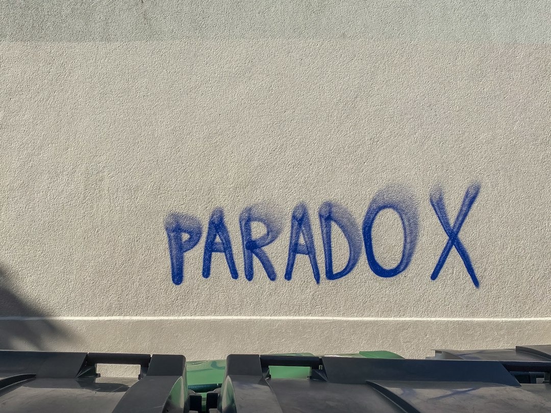 Blue spray-painted graffiti spelling 'PARADOX' on a light textured wall, with garbage/recycling bins at the bottom edge.