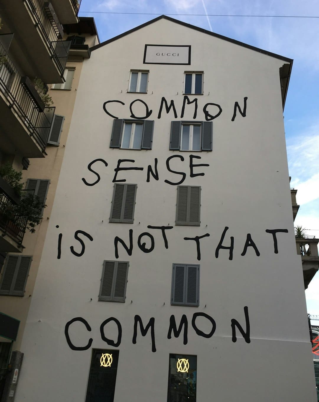 White building façade covered with large black graffiti text: 'COMMON SENSE IS NOT THAT COMMON', with a Gucci plaque near the roof and shuttered windows across the wall.
