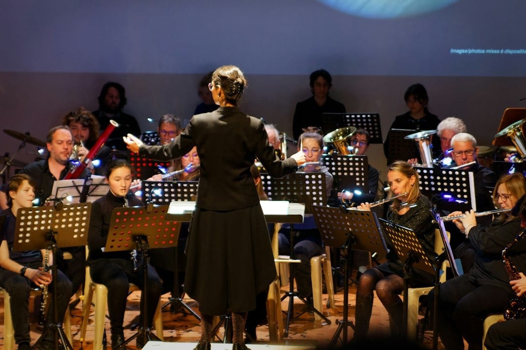 Conductor facing and directing a full orchestra on stage, musicians reading sheet music and playing instruments.